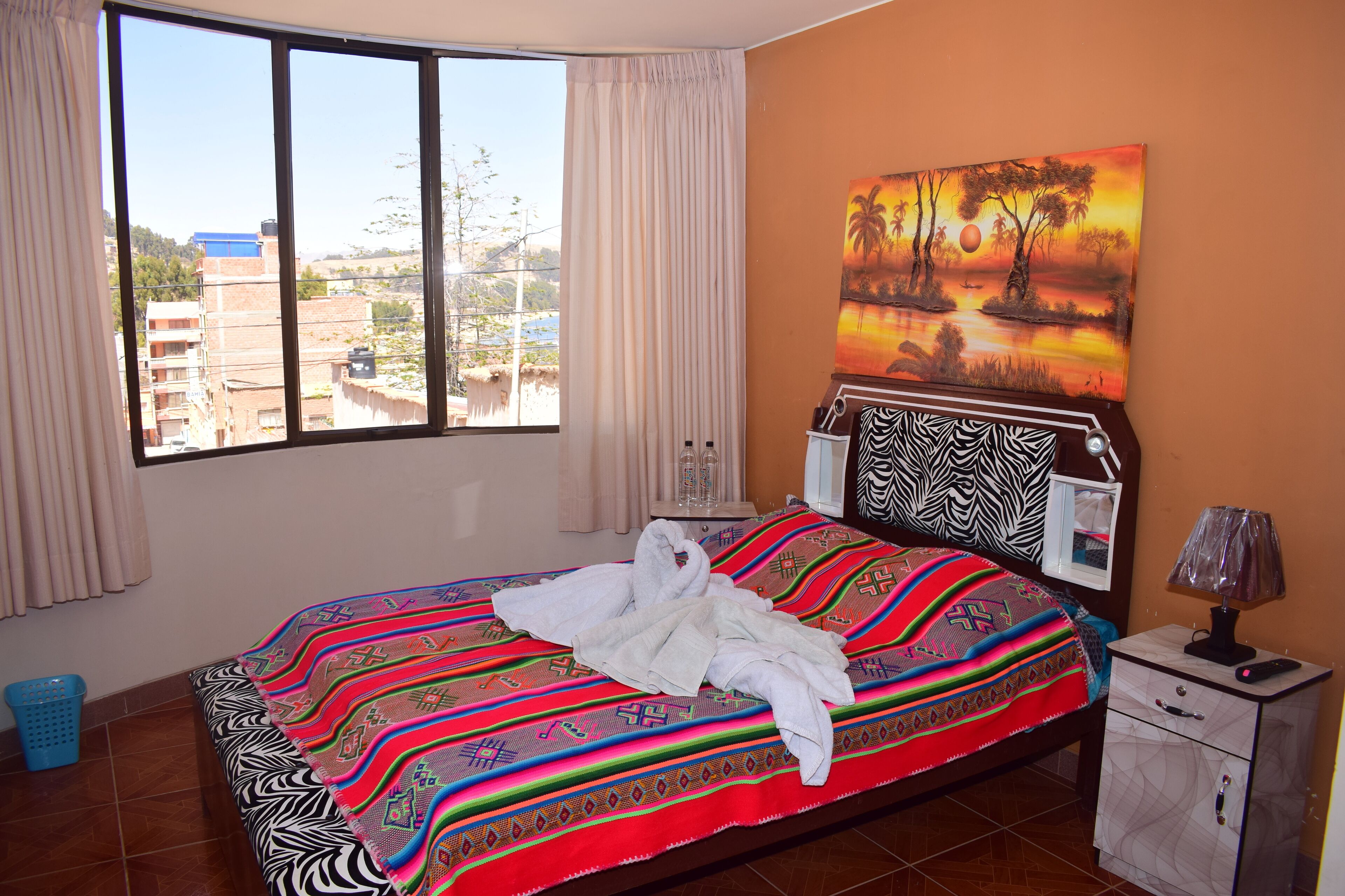 Quarto de hotel com uma cama grande, uma mesa de cabeceira, um abajur e uma pintura de um pôr do sol.