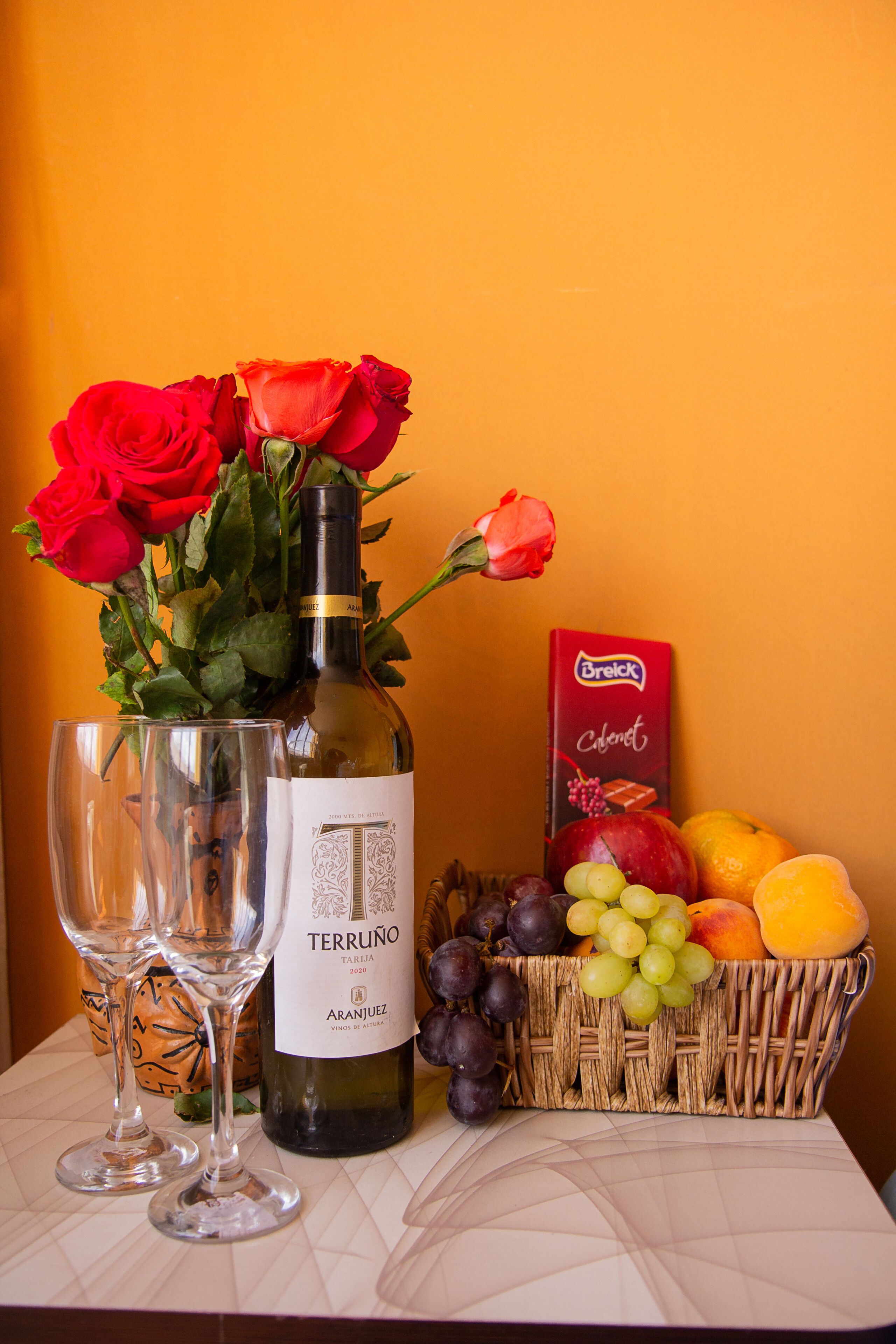 A table with a bottle of wine, two glasses, a basket of fruit, and a box of chocolates.
