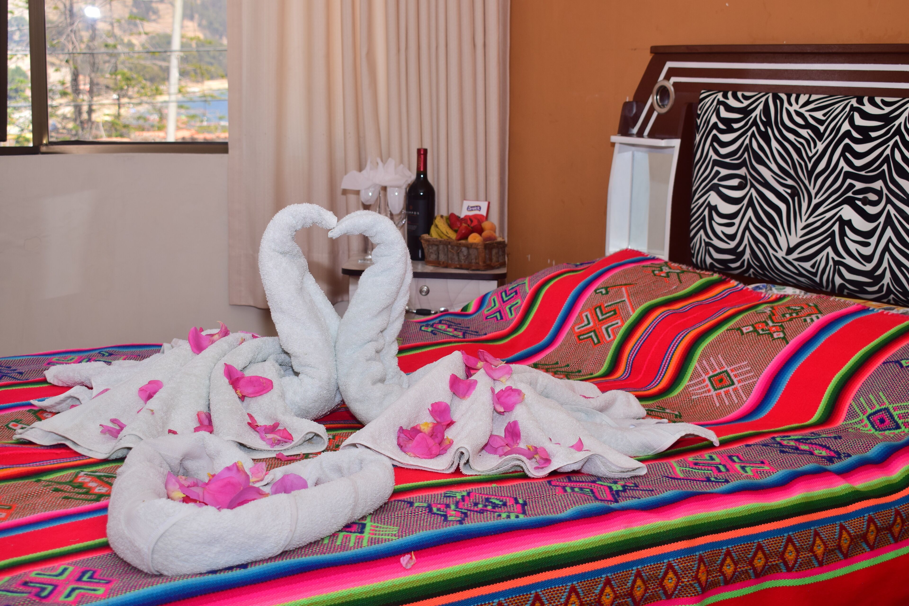 A hotel room with a bed featuring a colorful blanket, a decorative towel arrangement, a bottle of wine, and a fruit basket on the nightstand.