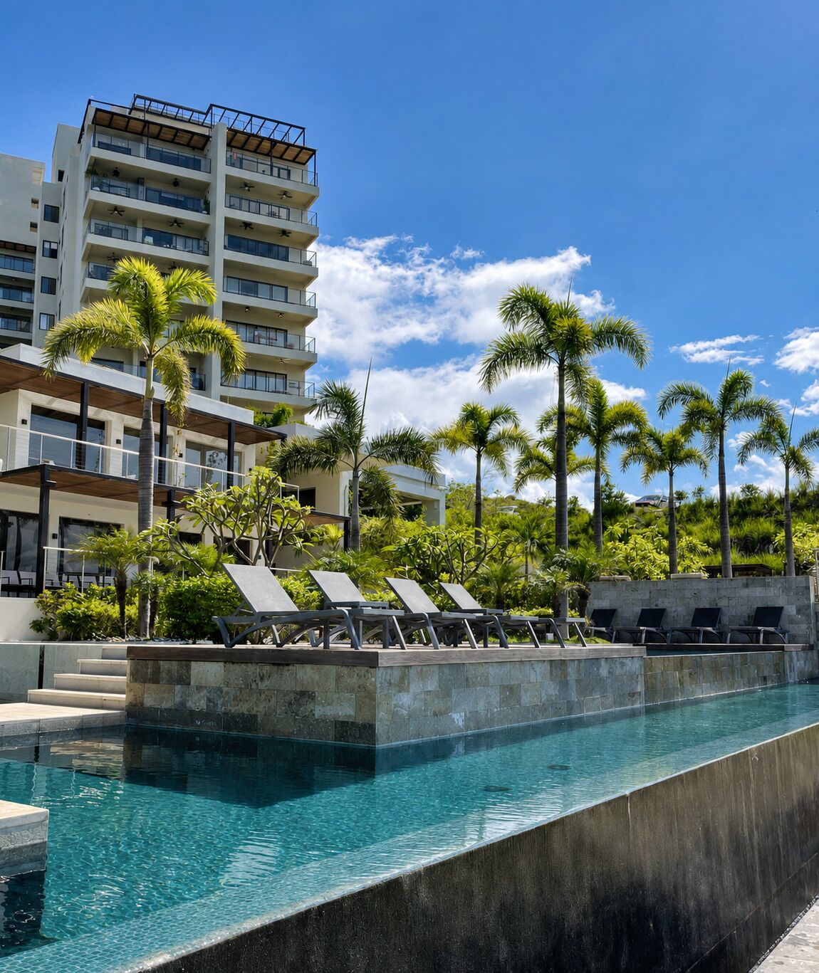 A swimming pool with lounge chairs, palm trees, and a multi-story building in the background.