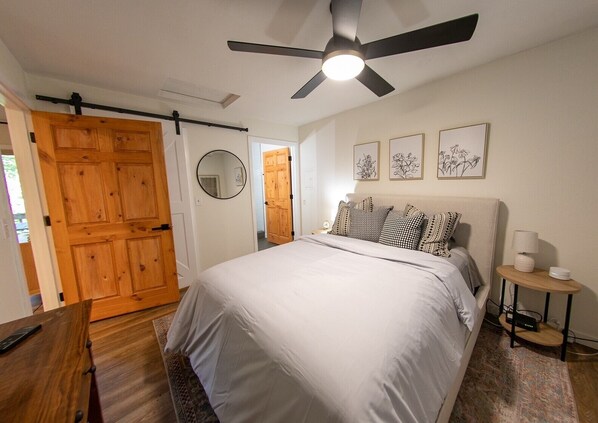 A bedroom with a bed, a ceiling fan, a bedside table with a lamp, and a wooden door.