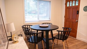 Un coin repas avec une table ronde et quatre chaises, une horloge et une fenêtre avec des stores.