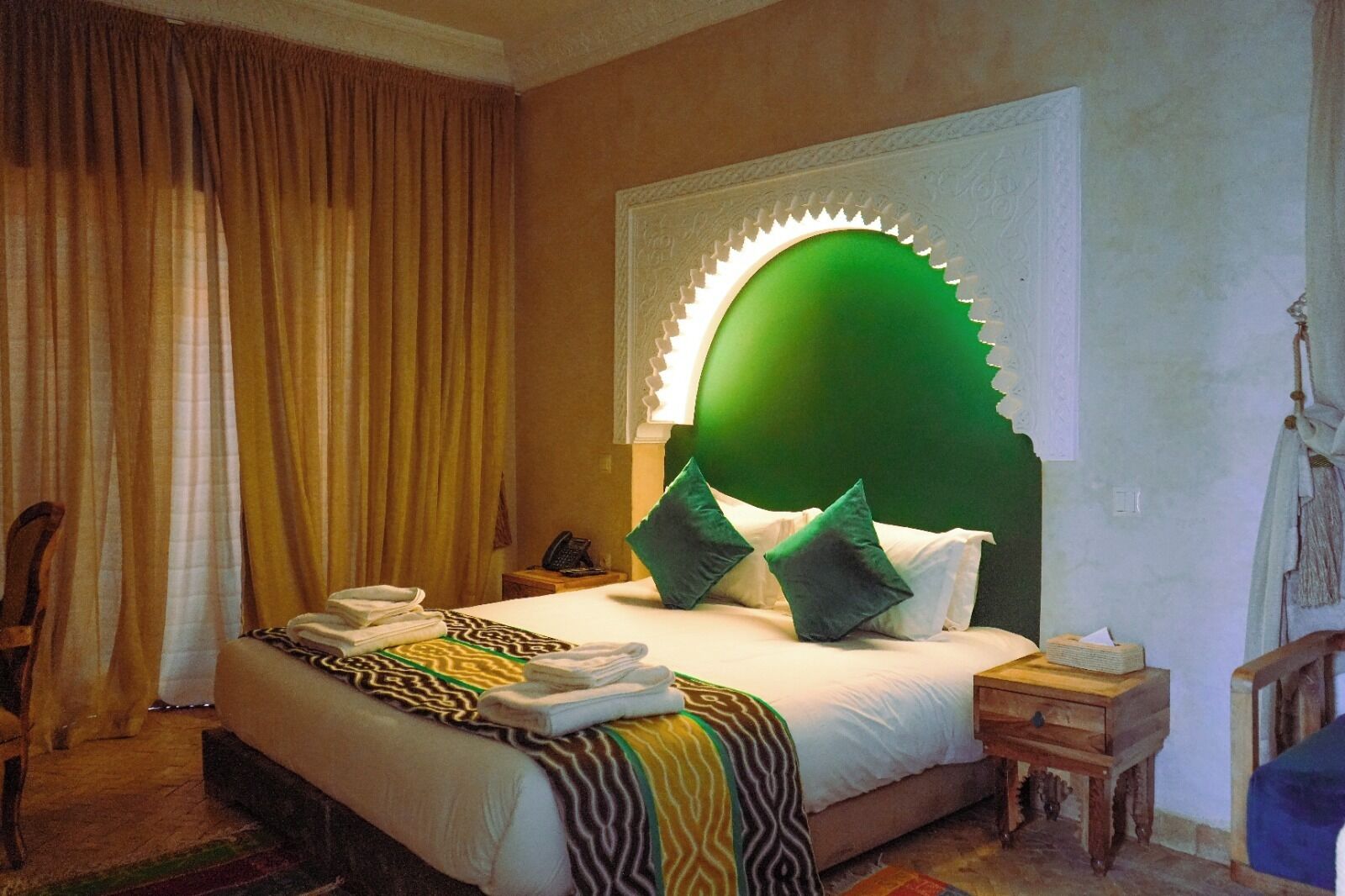 Una habitación de hotel con una cama grande, una mesita de noche, una silla y un ventanal con cortinas.