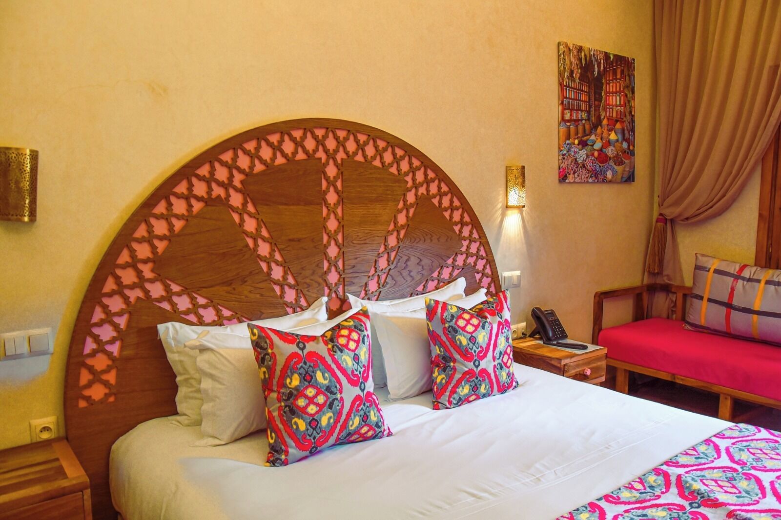 Quarto de hotel com uma cama grande, duas almofadas estampadas, uma cabeceira de madeira, uma mesa de cabeceira com telefone e uma cadeira vermelha.