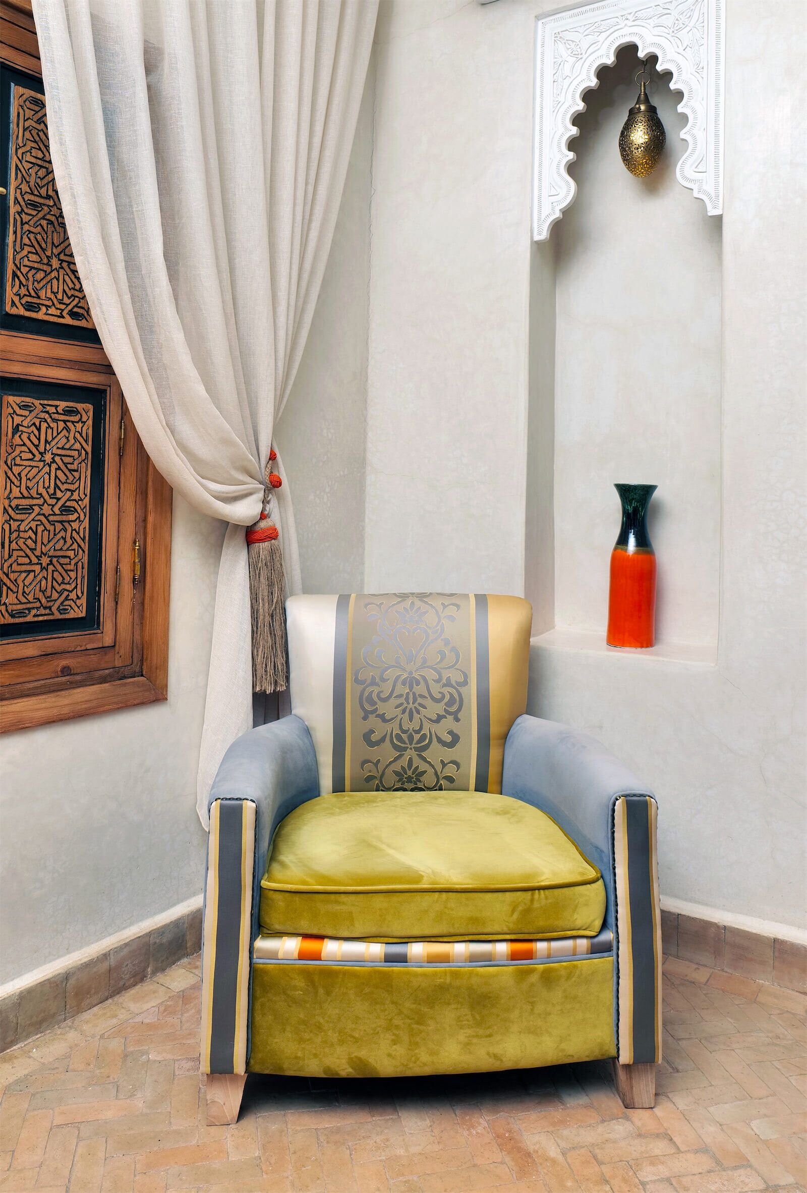 A corner of a room with a green upholstered chair, a patterned cushion, a hanging lamp, and a decorative wall piece.
