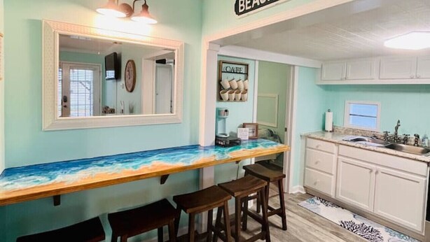 Una cocina con un letrero que dice 'BEACH HOUSE', taburetes de madera, una encimera con salpicadero decorativo y armarios blancos.