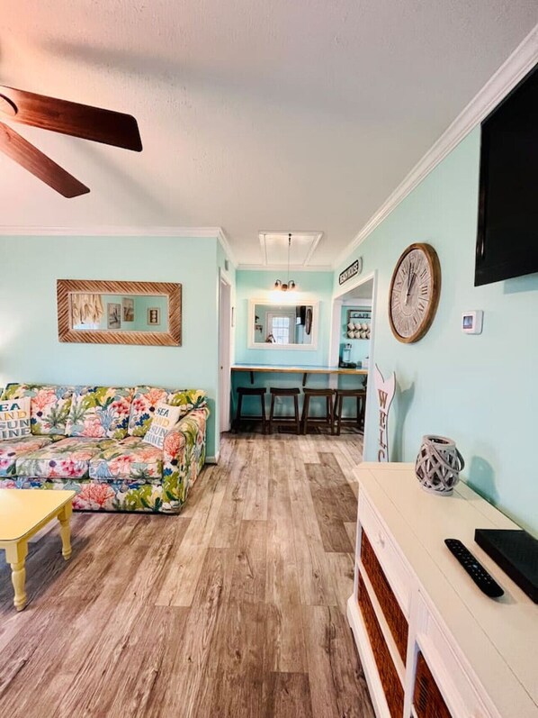 A living room with a floral sofa, wooden flooring, a ceiling fan, a TV, and a kitchen area in the background.