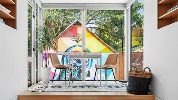 Un área de estar moderna al aire libre con una mesa, sillas, una planta en maceta y un mural colorido de fondo.