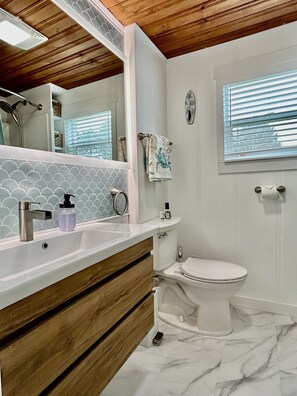 Un bagno con lavandino bianco, mobile contenitore in legno, uno specchio, un WC e una finestra con persiane.