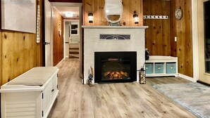 Een woonkamer met een open haard, houten wanden, een wit bakstenen haardmantel, een spiegel boven de haard en een blauwe opbergkast.