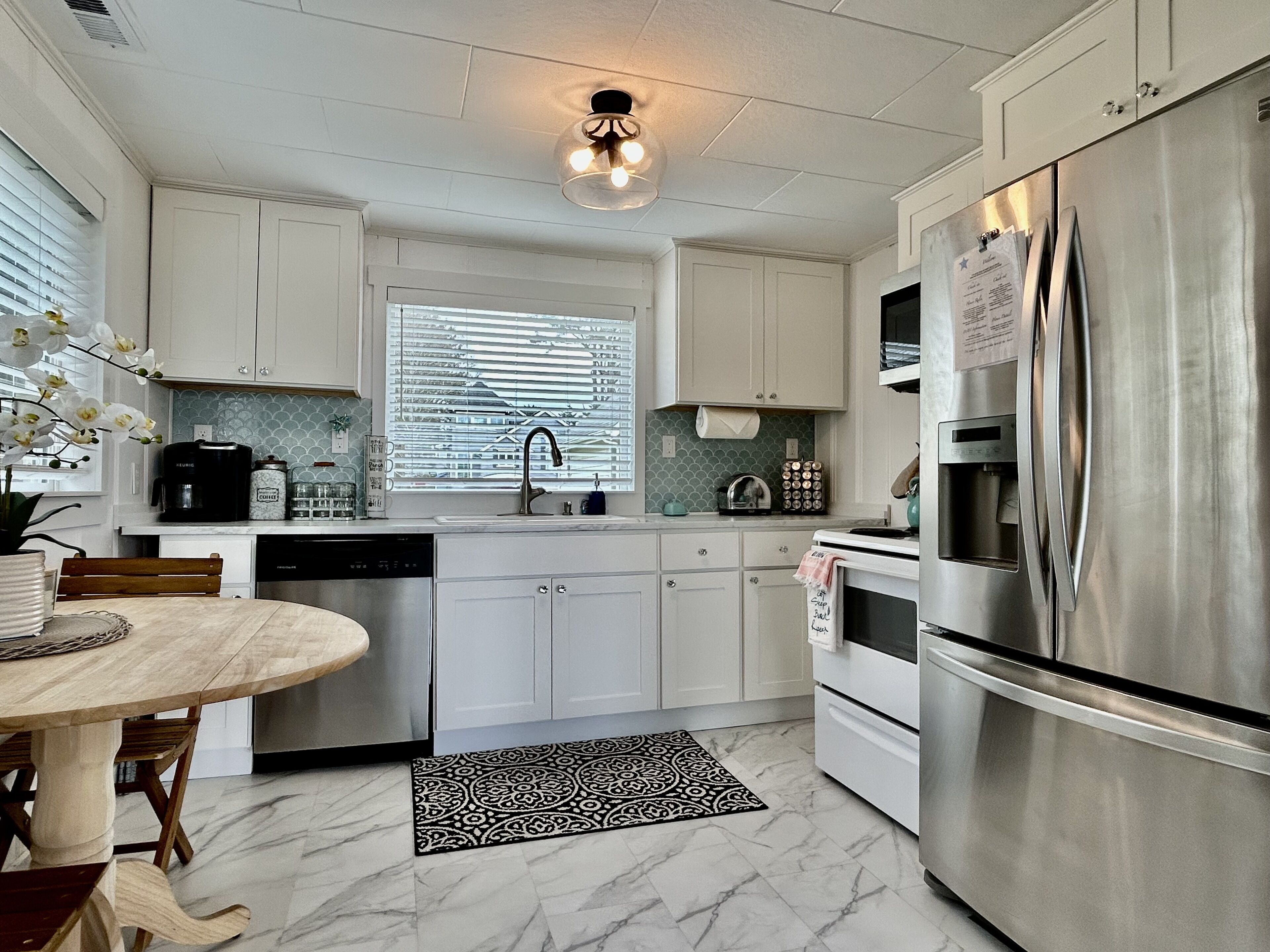 A modern kitchen with white cabinets, a stainless steel refrigerator, a coffee maker, and a window with blinds.
