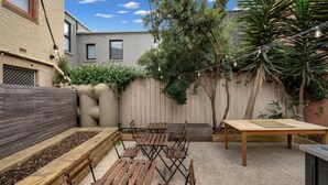 Un patio trasero con una mesa de madera y bancos, un pequeño jardín y un árbol.