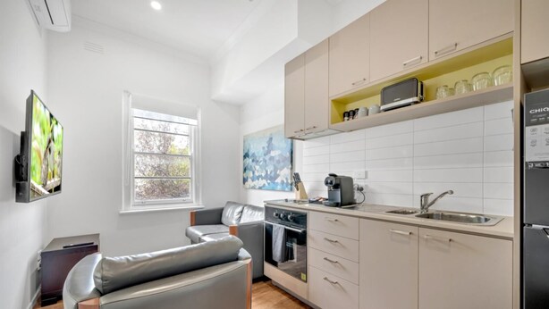 Cocina moderna con horno empotrado, microondas y lavabo. Hay un sofá gris y una ventana con vistas a árboles.