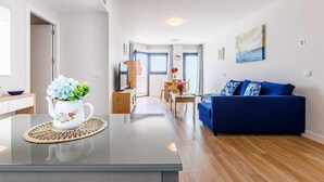 Een moderne woonkamer met een blauwe bank, een bijzettafeltje en een keukenhoek met een witte vaas met bloemen.
