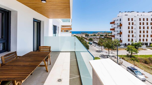 Een modern balkon met een houten tafel en stoelen, met uitzicht op een kustgebied met gebouwen en een heldere lucht.