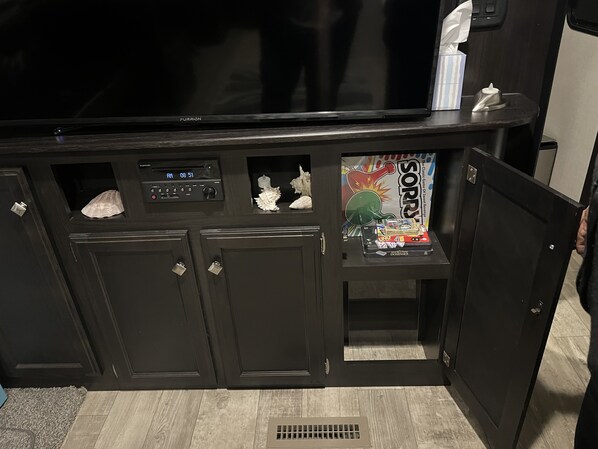 A room with a large flat-screen TV, a console with a clock, and a shelf with a game box and other items.