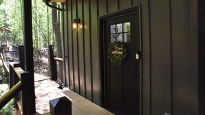 A wooden deck with a bench, a black door with a 'welcome' sign, and a lantern on the wall. - Luxury Treehouse 20 miles from asheville.  (Saluda)
