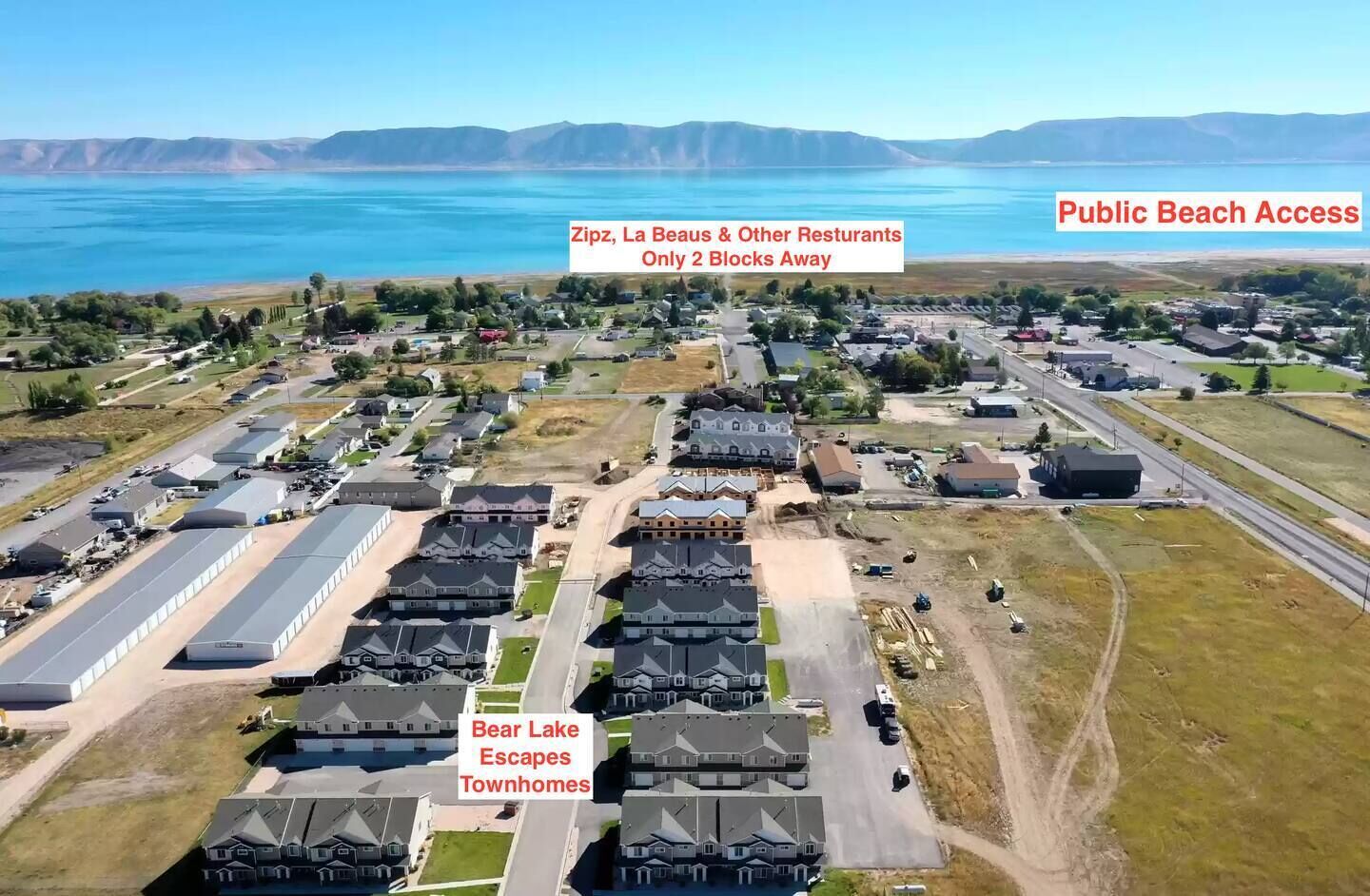 Luchtbeeld van een woongebied met huizen, een weg en een groot waterlichaam op de achtergrond. Tekstoverlays geven de nabijheid aan van Zipz, La Beaus & andere restaurants en toegang tot het openbare strand.