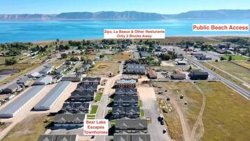 Luchtbeeld van een woongebied met huizen, een weg en een groot waterlichaam op de achtergrond. Tekstoverlays geven de nabijheid aan van Zipz, La Beaus & andere restaurants en toegang tot het openbare strand.