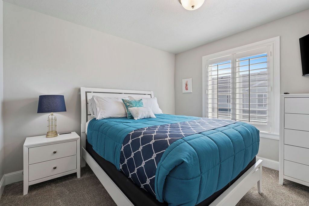 A bedroom with a bed, a nightstand, a lamp, a window with blinds, and a TV on the wall.
