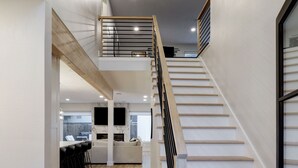 A modern interior with a staircase leading to a second floor, wooden flooring, and a kitchen area with bar stools. - Modern Farmhouse • Chef Kitchen • GameRoom • 77" OLED • BBQ Grill  (Katy)
