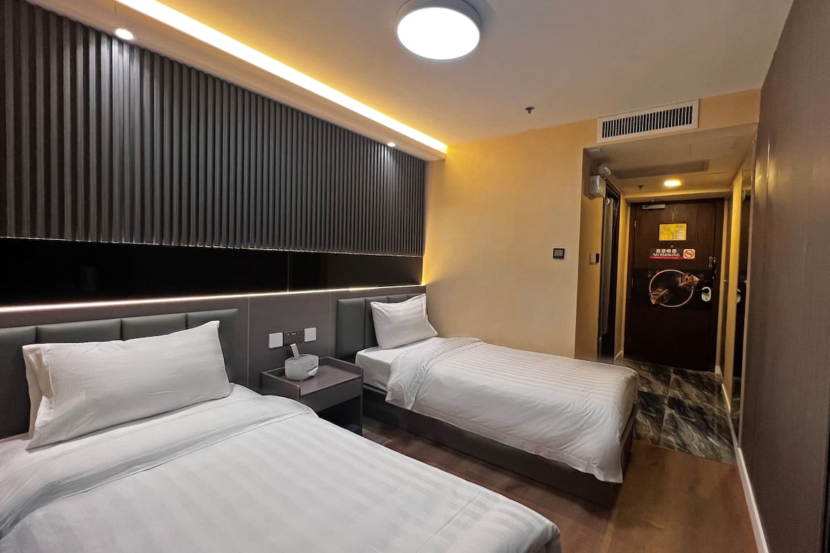 A hotel room with two beds, a bedside table with a lamp, a wall-mounted TV, and a door with a 'No Smoking' sign.