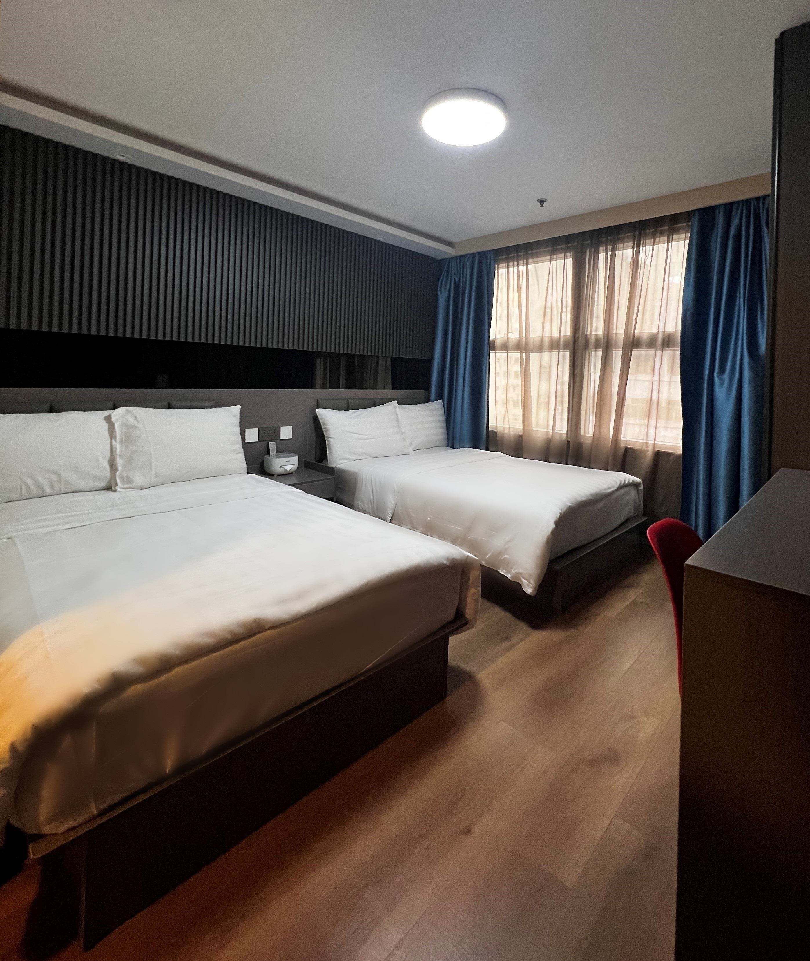 A hotel room with two beds, a desk, a chair, and a window with curtains.