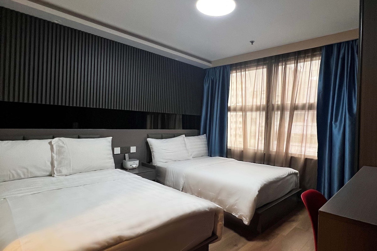 A hotel room with two beds, a desk, a chair, and a window with curtains.