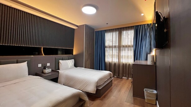 A hotel room with two beds, a TV mounted on the wall, a bedside table with a lamp, and a window with curtains. - Hennessy Hotel (Hong Kong)