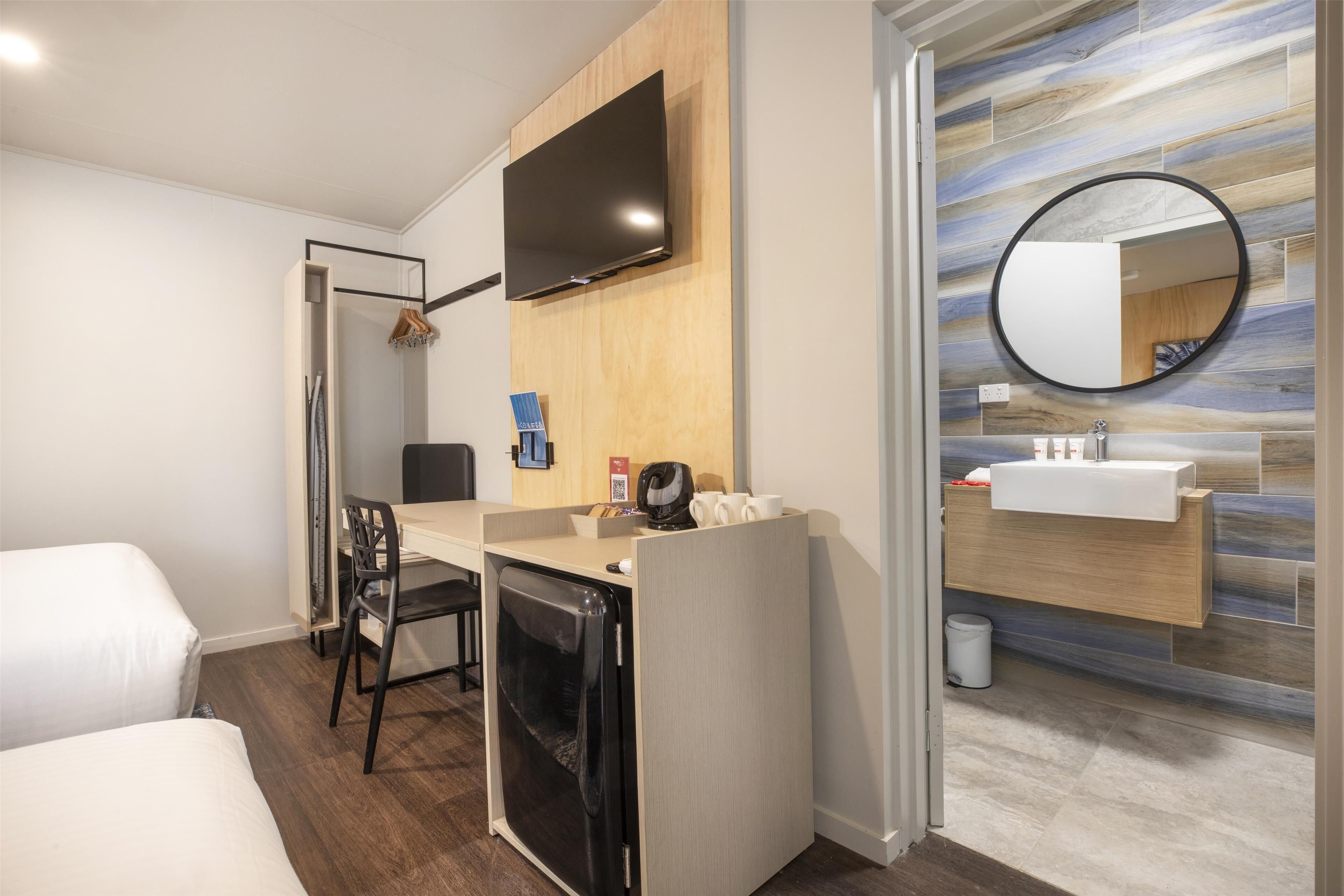 Ein Hotelzimmer mit Schreibtisch, Stuhl und einer kleinen Küchenzeile. Ein Fernseher ist an der Wand angebracht und über dem Waschbecken befindet sich ein Spiegel.