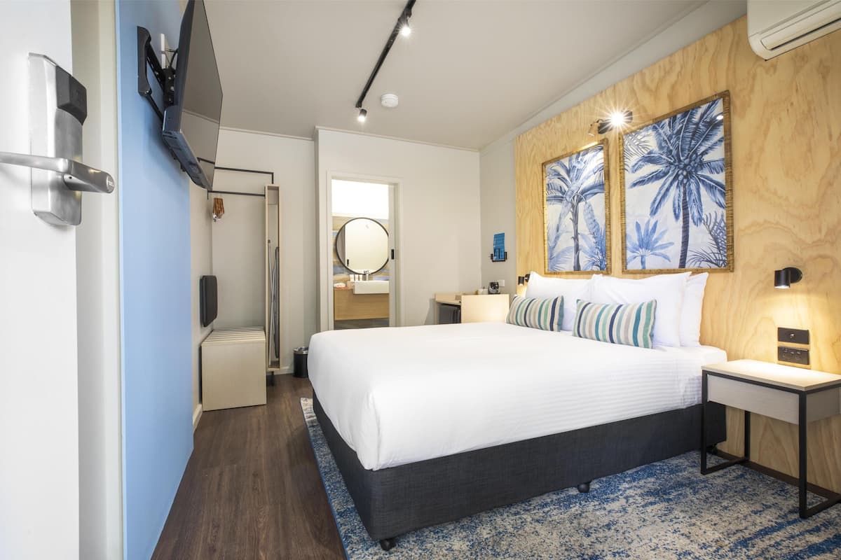 Habitación de hotel moderna con una cama grande, un televisor de pantalla plana en la pared, un panel de madera decorativo con obras de arte, una mesita de noche con lámpara y un baño visible a través de una puerta abierta.