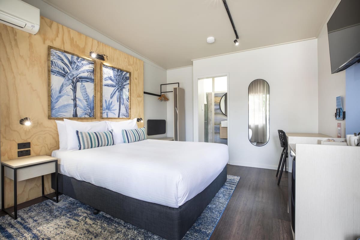 Habitación de hotel moderna con una cama grande, un escritorio y un espejo. Hay un cuadro en la pared y una puerta que da al baño.