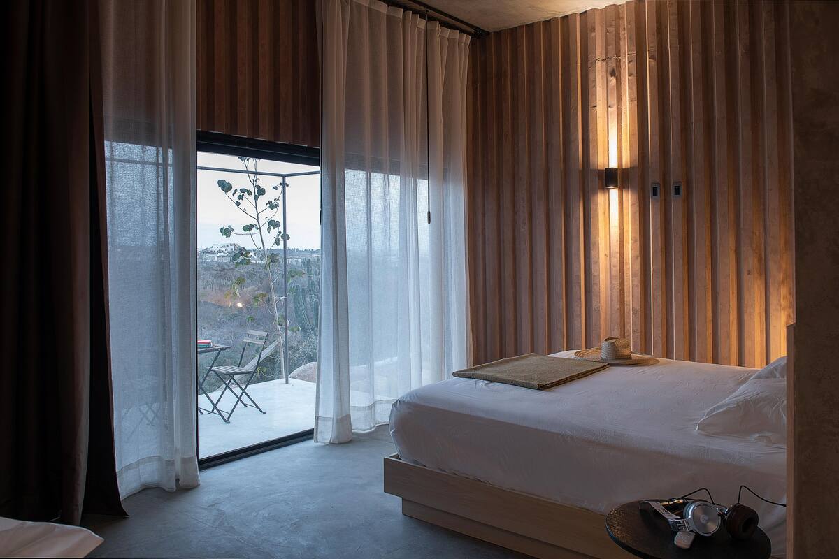 A bedroom with a large bed, a view through the window, and a table with a plant.
