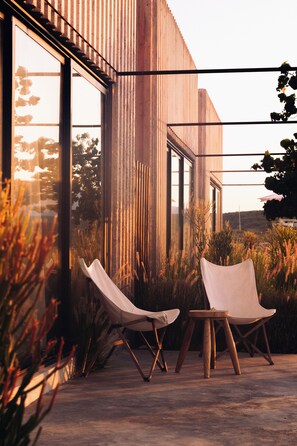 Un espacio exterior moderno con dos sillones reclinables y piso de concreto, rodeado de pastos altos y un edificio con amplios ventanales.