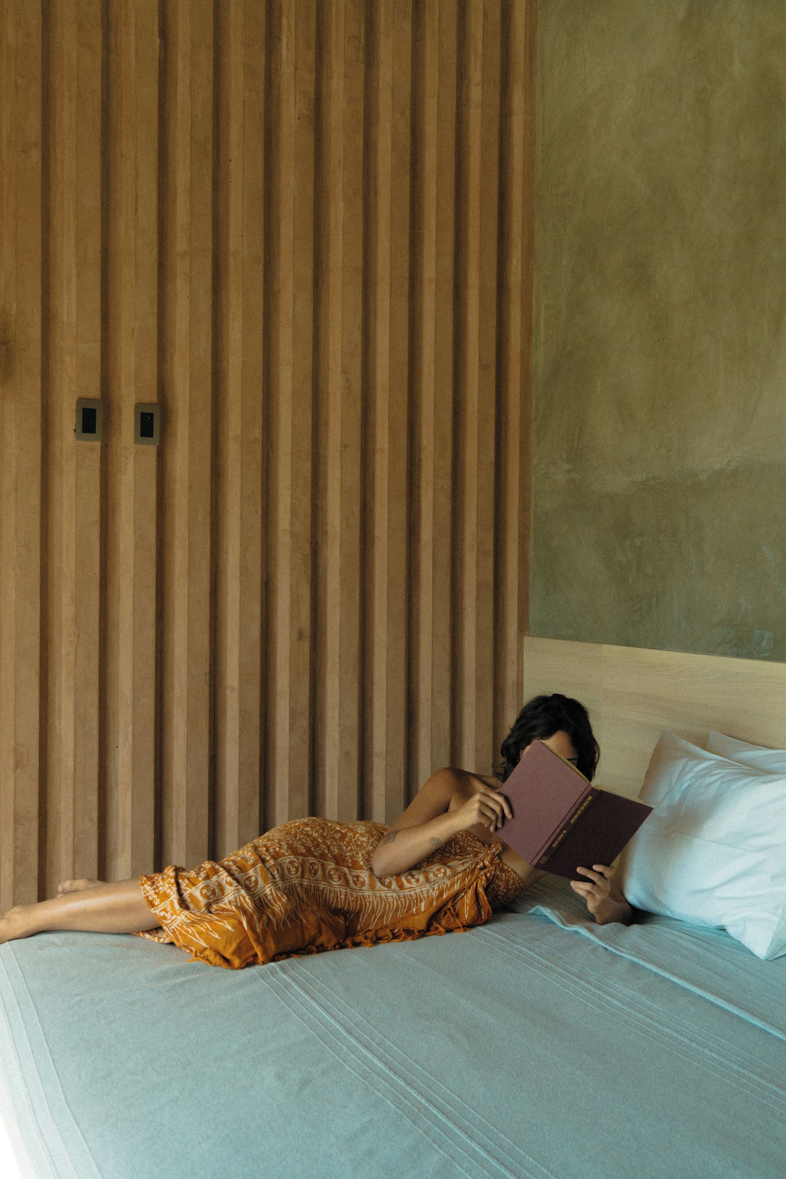 Eine Person liegt mit einem Buch auf einem Bett, in einem Zimmer mit Holzvertäfelung und einer schlichten Wand.
