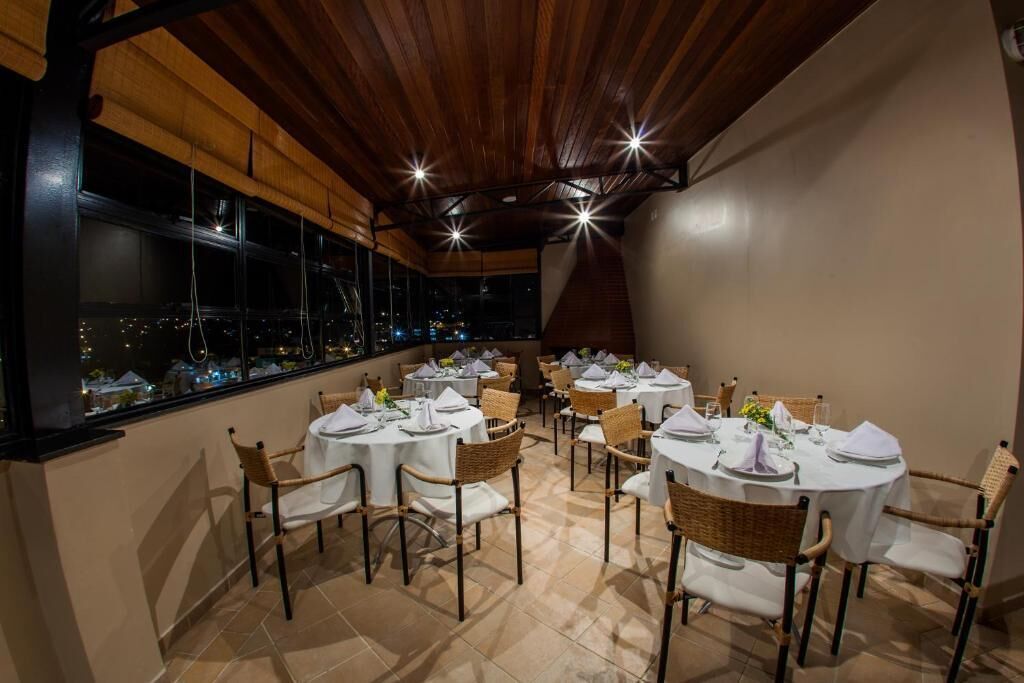 Un comedor con mesas redondas dispuestas para cuatro comensales, cada una con mantel blanco, sillas de mimbre y vistas a la ciudad a través de amplias ventanas.
