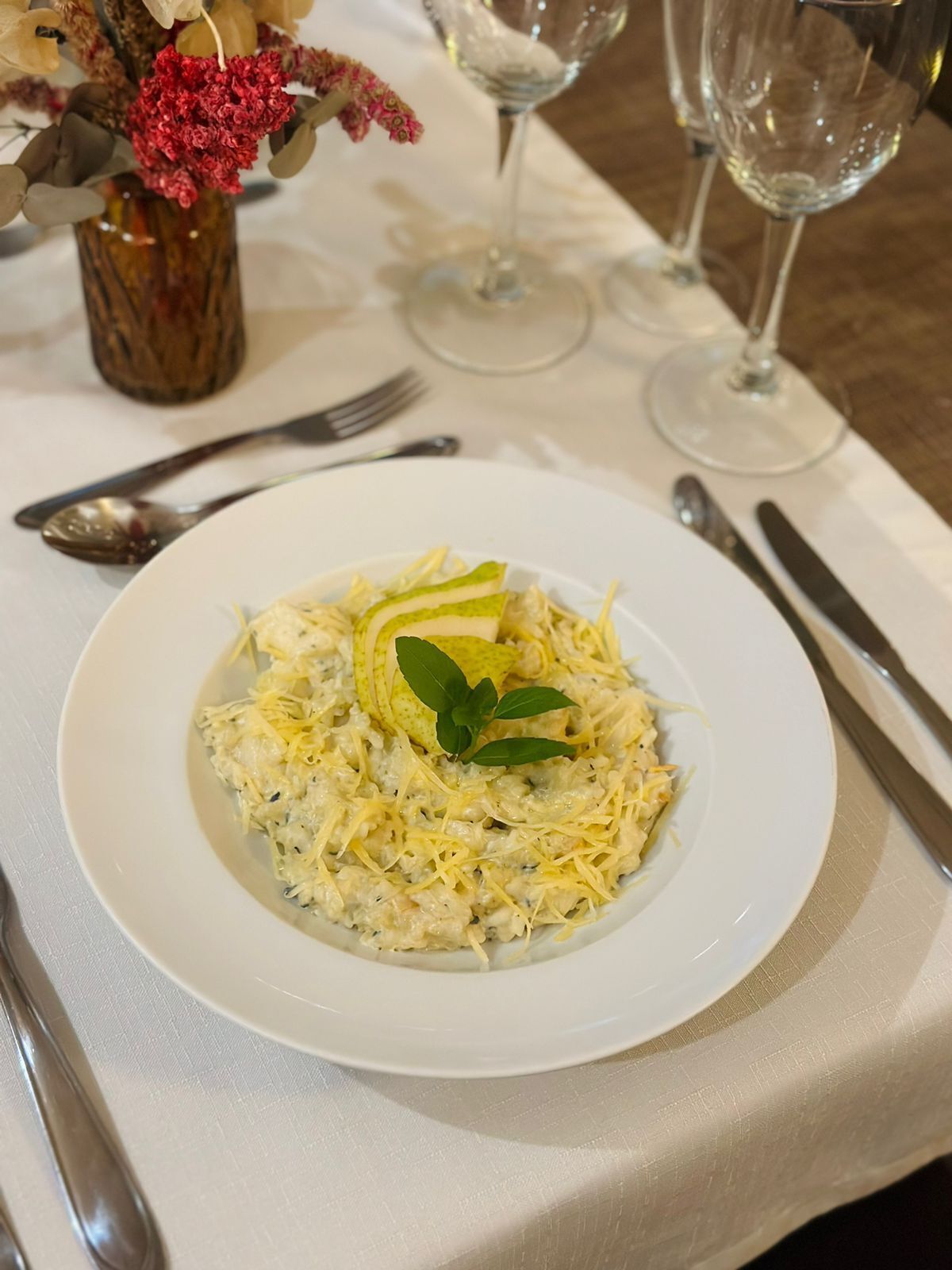 A plate of food with shredded cheese and a garnish, set on a table with a vase of flowers and wine glasses.