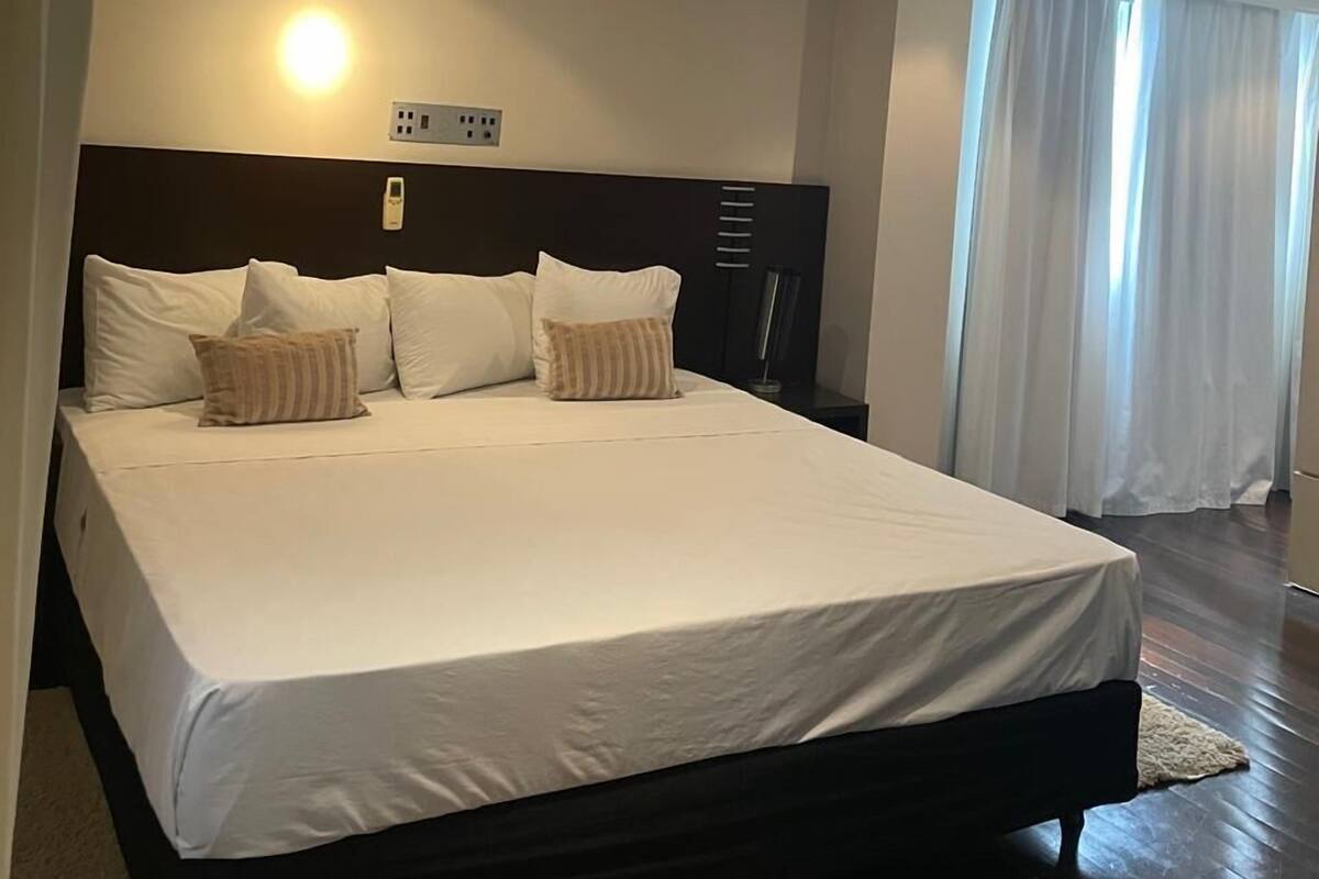 Una cama grande con ropa blanca, un cabecero oscuro y una mesita de noche con una lámpara.