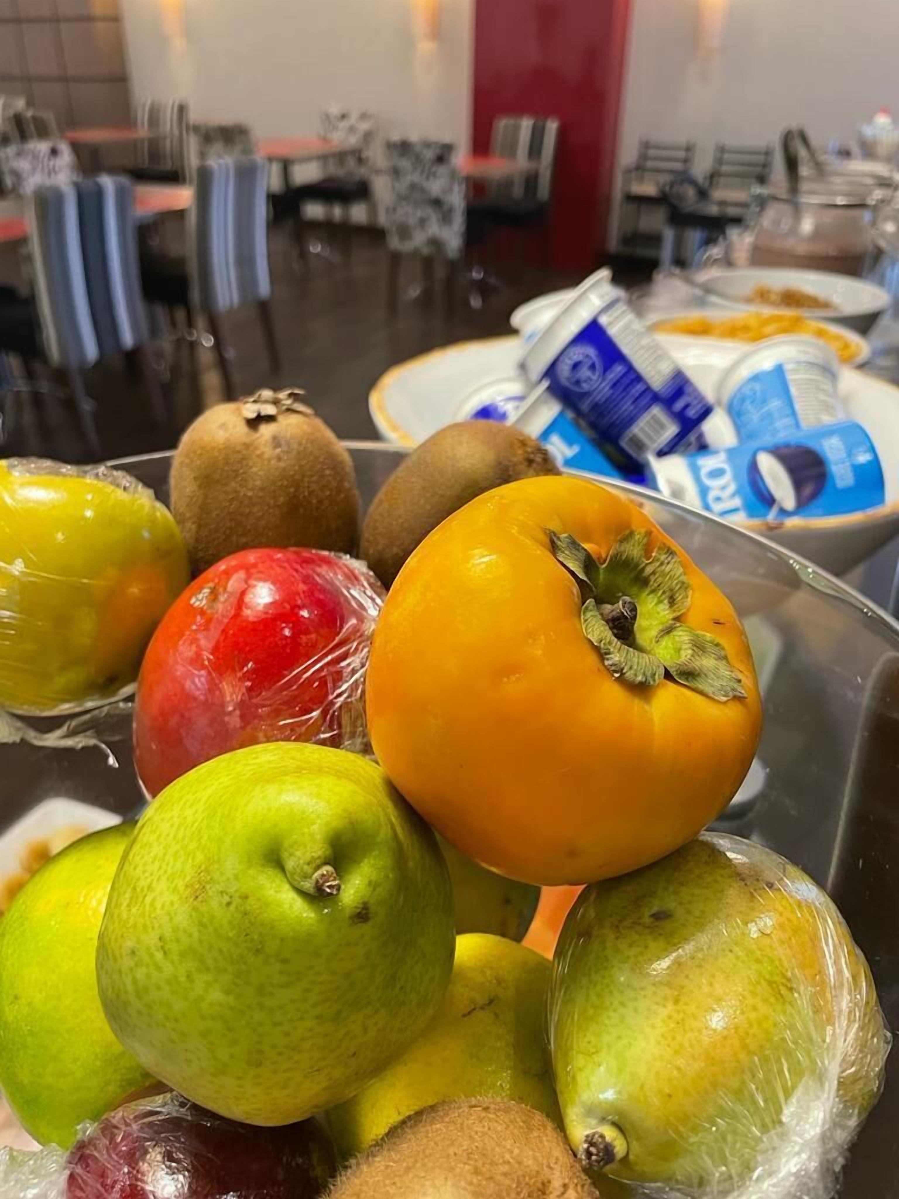 A variety of fruits including apples, kiwis, and oranges on a table in a dining area with chairs and tables in the background.