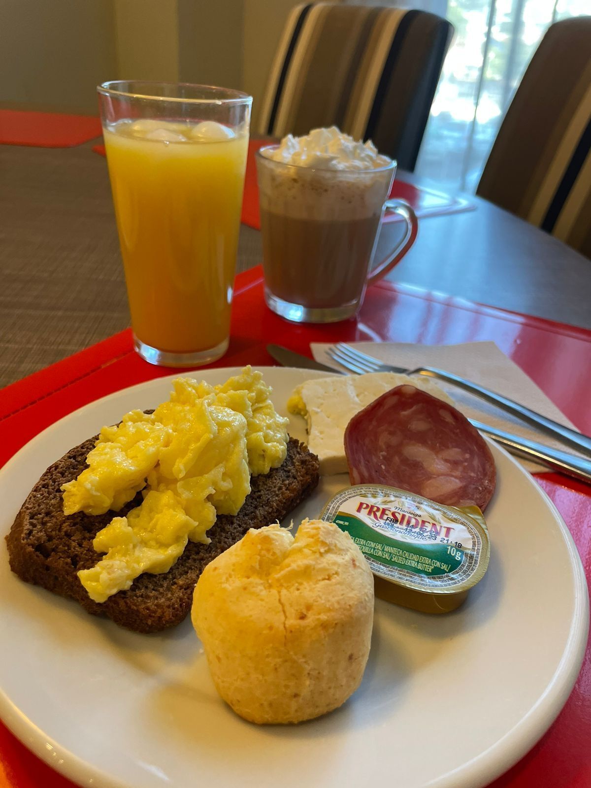 A breakfast plate with scrambled eggs, a biscuit, a slice of salami, and a container of yogurt, along with a glass of orange juice and a cup of coffee.