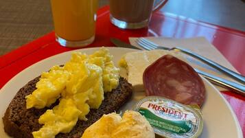 A breakfast plate with scrambled eggs, a biscuit, a slice of salami, and a container of yogurt, along with a glass of orange juice and a cup of coffee.