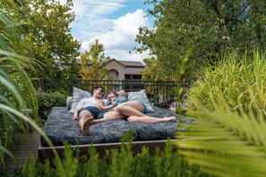 A couple lying on a bed with a patterned mattress, surrounded by greenery and a fence, with a building in the background.