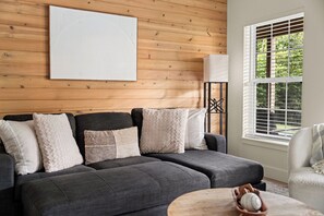A modern living room with a dark grey sofa, multiple cushions, a white abstract wall art piece, a side table with a decorative item, and a window with blinds.