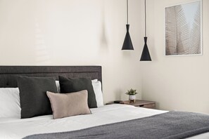 A neatly made bed with a grey headboard, a bedside table with a plant, and a wall-mounted artwork.
