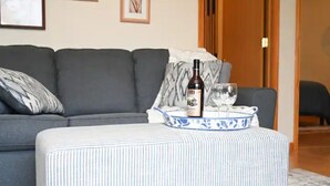 Una sala de estar con un sofá gris, un reposapiés a rayas, una bandeja azul y blanca con una botella y vasos, y un cuadro en la pared.