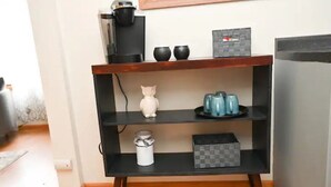 Una cocina con un mueble negro que alberga una cafetera, dos tazas negras, una figurita de gato pequeño, un recipiente blanco y una taza azul.