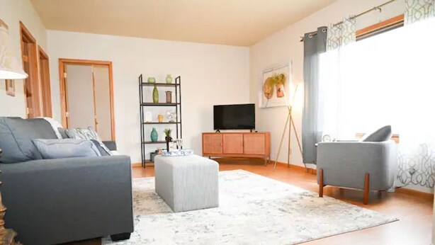 Una sala de estar con un sofá gris, un reposapiés gris, un mueble de madera y un televisor.
