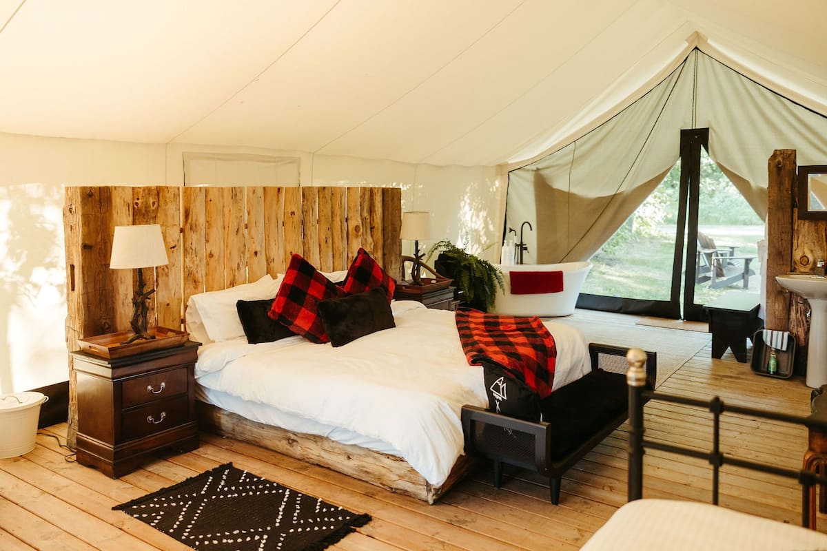 Ein gemütliches Schlafzimmer mit einem hölzernen Kopfteil, einem Bett mit roten und schwarzen Kissen, einem Nachttisch mit einer Lampe und Blick ins Freie durch ein zeltartiges Fenster.
