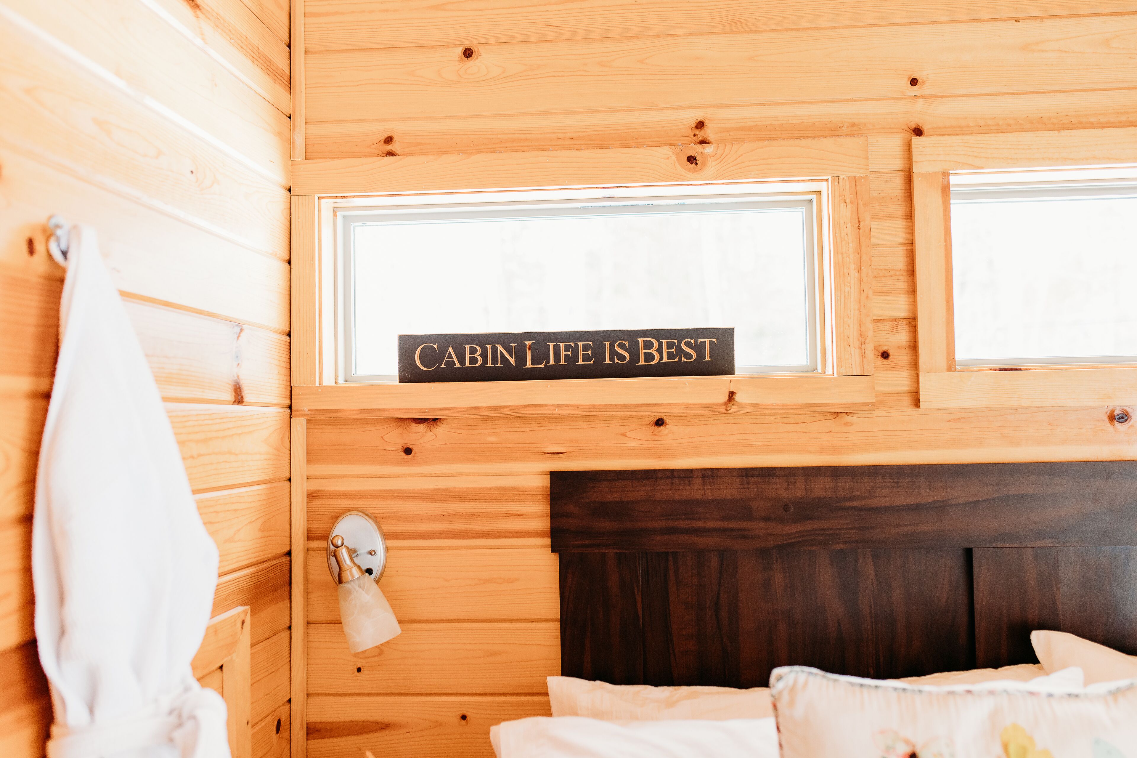 A cabin interior with wooden walls, a bed with white bedding, and a sign above the window stating "CABIN LIFE IS BEST".