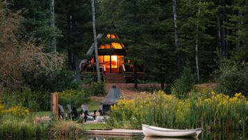 A cabin with a wooden deck, surrounded by a forest and a body of water.